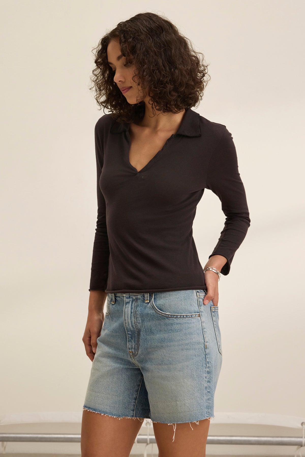 A person with curly hair stands sideways, hands in pockets, wearing a REED POLO by Velvet by Graham & Spencer and denim shorts against a plain background, reminiscent of early collections with its timeless appeal.-38928814112961