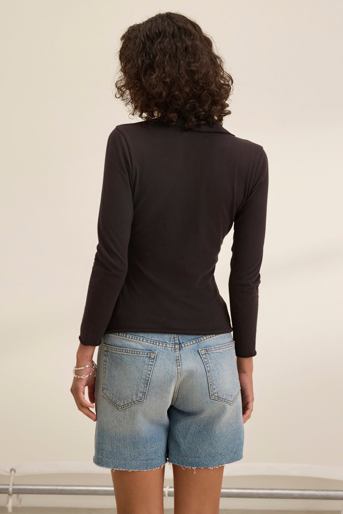 An individual with curly hair embodies timeless elegance, wearing the Velvet by Graham & Spencer REED POLO. Standing indoors against a light background, the black long-sleeve top and denim shorts nod to classic fashion simplicity.-38928814080193
