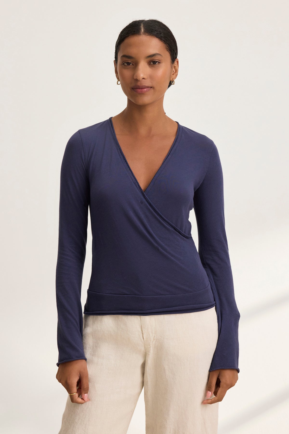 A person wearing the TANIA TEE, a classic long-sleeve navy wrap top by Velvet by Graham & Spencer, and beige pants stands against a plain light background, looking at the camera.-38928814768321