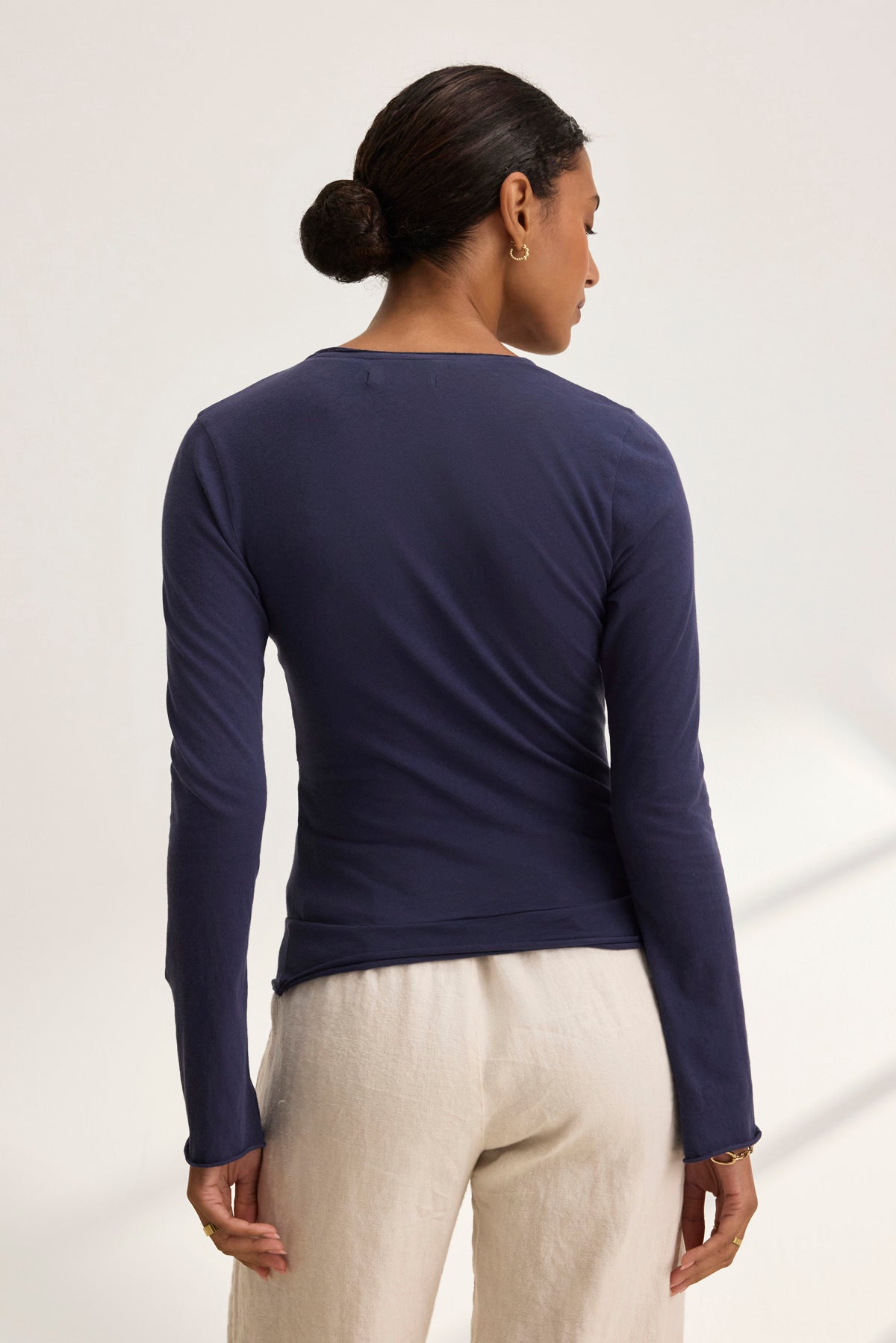 The individual, hair tied in a classic bun, wears the Velvet by Graham & Spencer TANIA TEE in dark blue with light-colored pants against a plain backdrop, exuding timeless style.-38928814604481