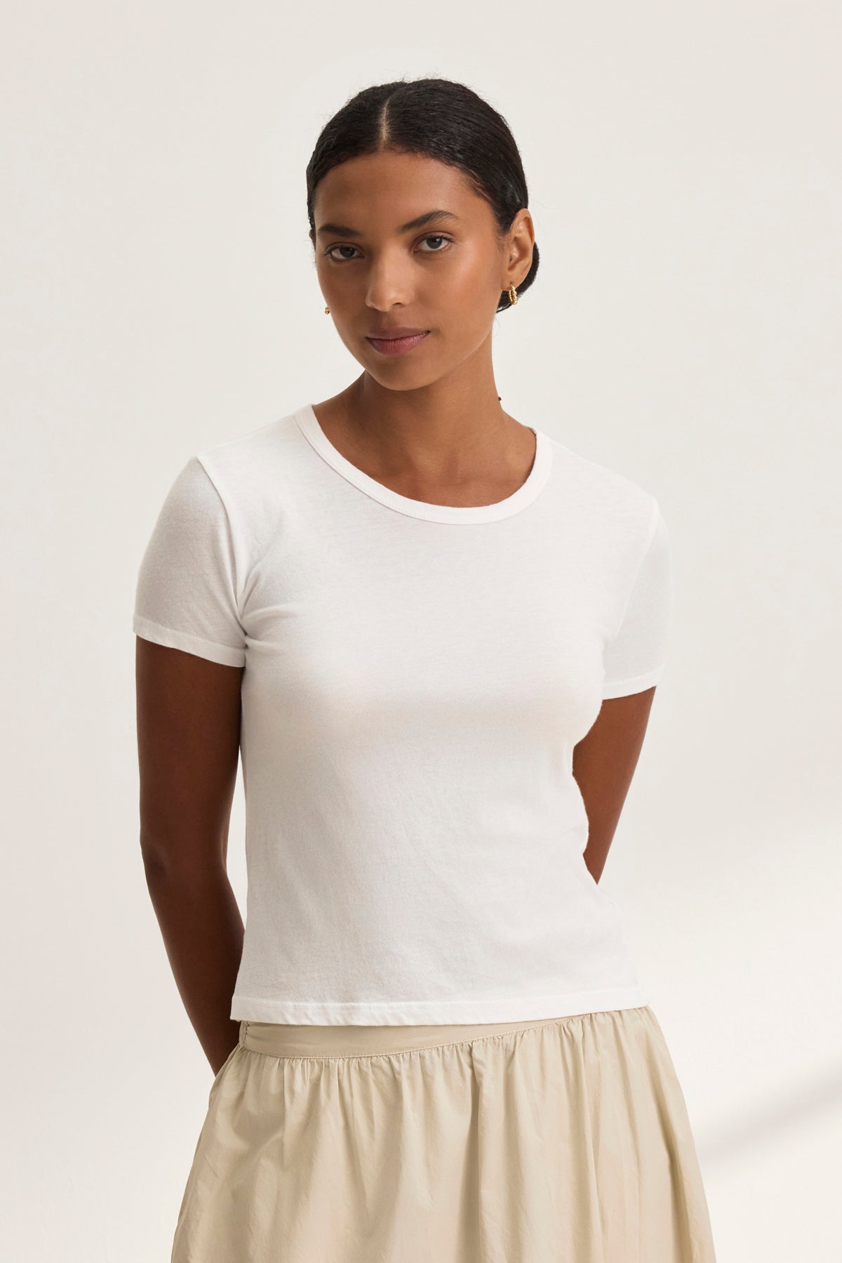 A person with short, dark hair wearing the WHITNEY TEE by Velvet by Graham & Spencer and a beige skirt embodies a classic, timeless style against a plain background.-38928815521985