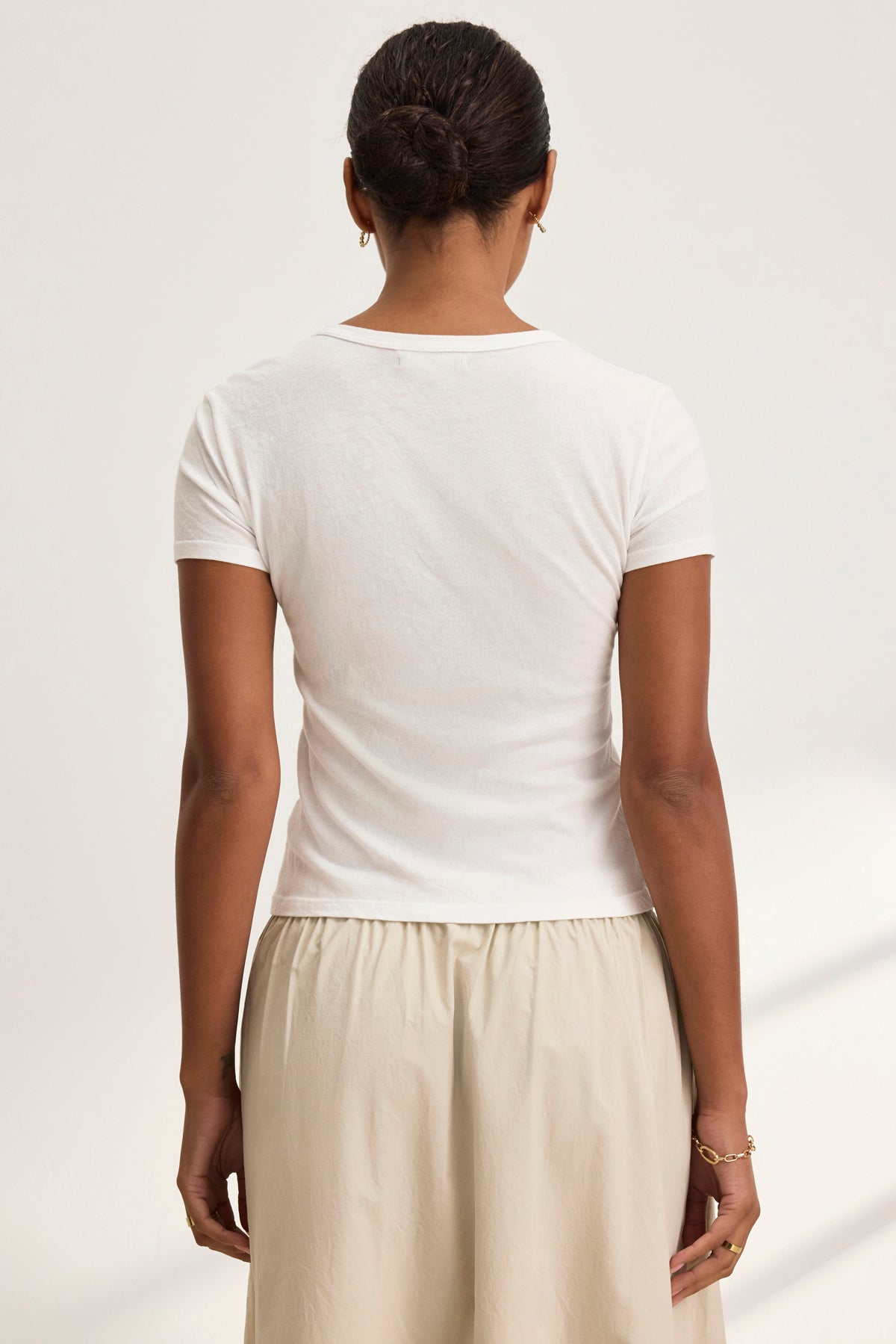 A person with dark hair in a bun, wearing the WHITNEY TEE by Velvet by Graham & Spencer paired with a beige skirt, stands with their back to the camera, embodying classic timeless elegance.-38928815456449