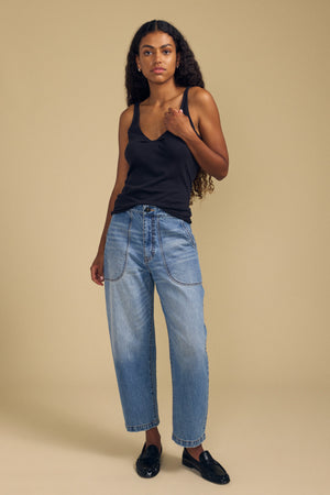 Woman with long curly hair stands against a beige background, wearing the KIM TANK TOP by Velvet by Graham & Spencer—a black cotton tank featuring a scoop neckline—paired with light blue jeans and black loafers for an essential wardrobe look.