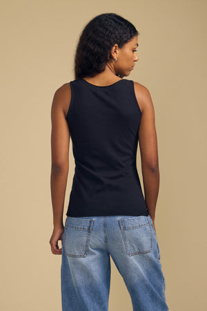A person stands facing away, wearing the KIM TANK TOP by Velvet by Graham & Spencer—a black cotton tank with a scoop neckline—paired with light blue jeans against a plain beige background, showcasing this true wardrobe essential.