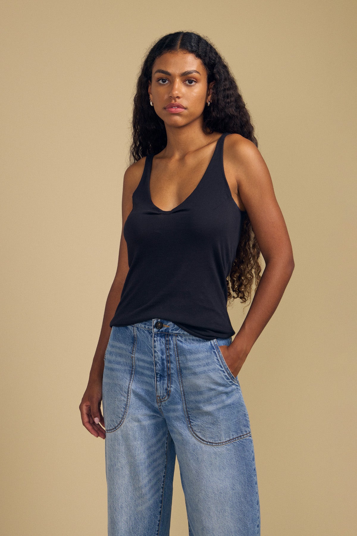 A woman with long curly hair wears the KIM TANK TOP by Velvet by Graham & Spencer, a black cotton tank featuring a scoop neckline, paired with blue jeans. She stands against a plain beige background with a neutral expression—making this piece a true wardrobe essential.