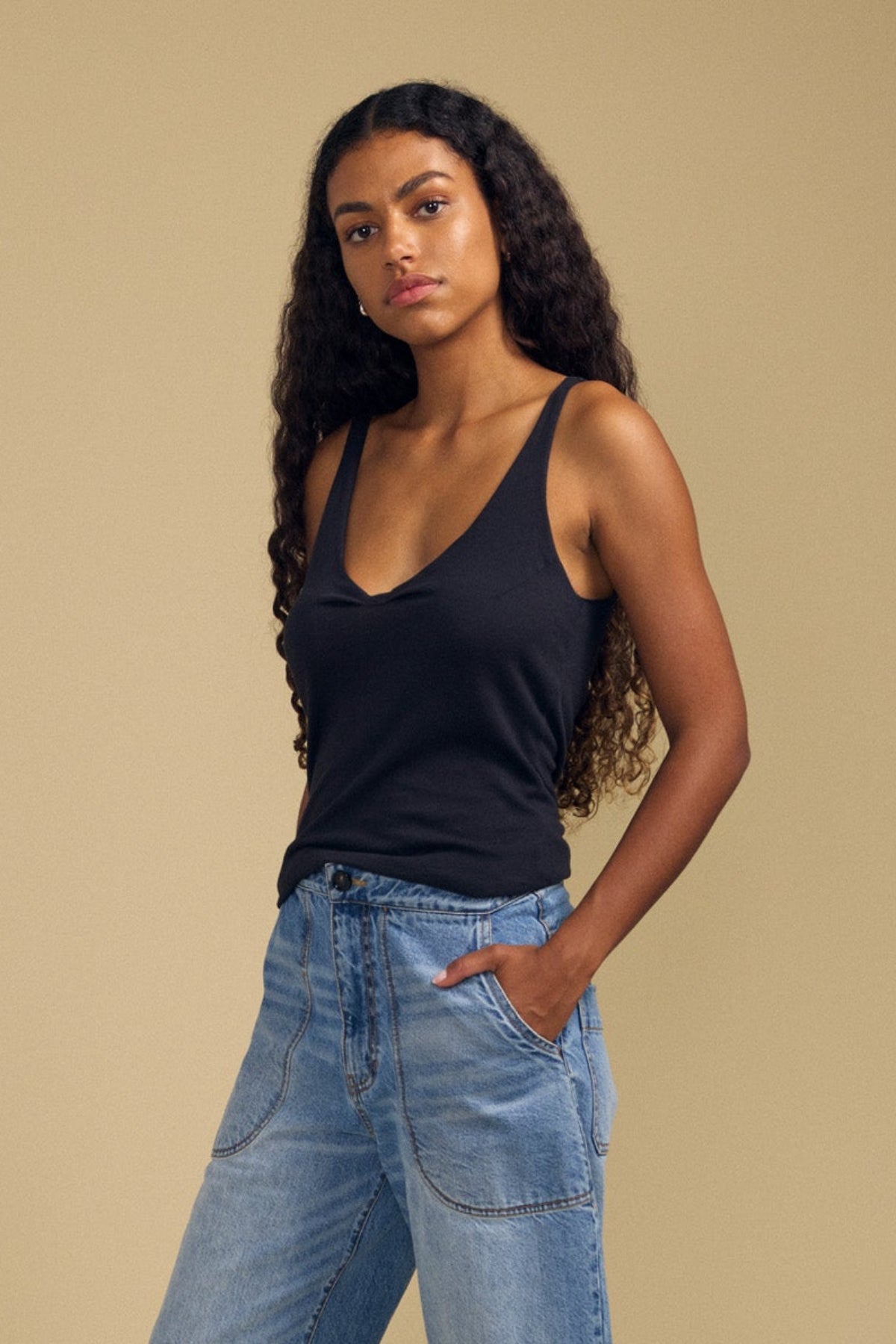 A woman with long curly hair wears the KIM TANK TOP by Velvet by Graham & Spencer—a black cotton tank featuring a scoop neckline—paired with light blue jeans as she stands against a beige background with her hands in her pockets, showcasing a true wardrobe essential.
