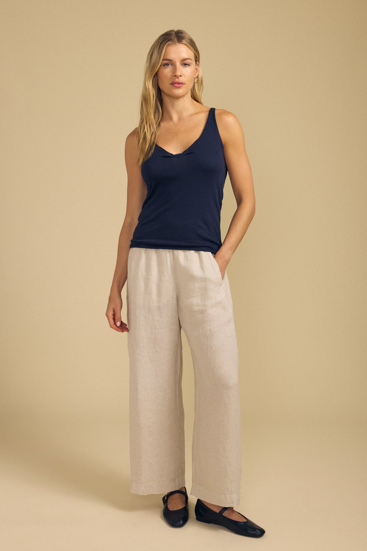 A woman stands against a beige background wearing the KIM TANK TOP by Velvet by Graham & Spencer—a navy, sleeveless scoop-neck top—paired with wide-leg beige linen pants and black flats. She has one hand in her pocket.
