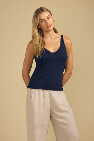 A person stands against a beige background, wearing the KIM TANK TOP by Velvet by Graham & Spencer in navy blue cotton with a scoop neckline, paired with loose, light-colored pants and their arms relaxed at their sides.