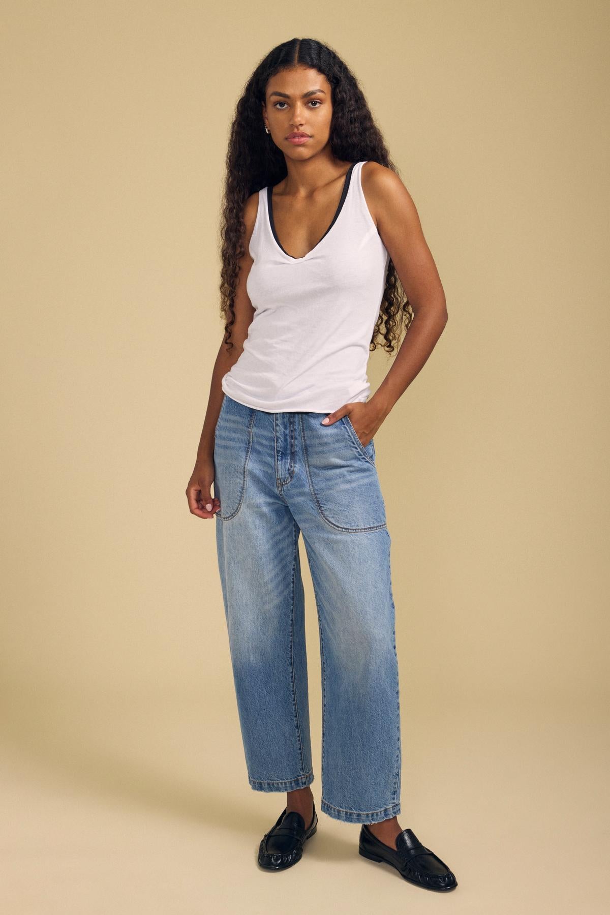A woman stands against a beige background wearing the KIM TANK TOP by Velvet by Graham & Spencer—a white cotton tank top—paired with loose blue jeans featuring pleat detailing and black loafers, with one hand in her pocket and the other relaxed at her side.-42125155270849