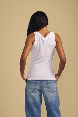 A person with long curly hair stands facing away against a plain beige background, wearing the KIM TANK TOP by Velvet by Graham & Spencer—a sleeveless white cotton essential paired effortlessly with blue jeans.