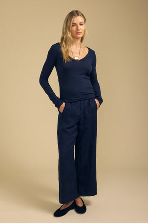 A woman stands against a beige background wearing the NOURA TEE by Velvet by Graham & Spencer—a navy long sleeve cotton jersey with a scoop V-neckline—paired with wide-leg navy pants, black shoes, and a pendant necklace.