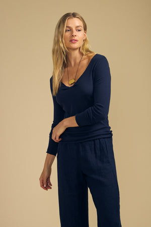 A person with long blonde hair wears the NOURA TEE by Velvet by Graham & Spencer—a navy cotton jersey top featuring a scoop V-neckline and long sleeves—paired with navy pants and a necklace with a tan pendant, standing against a beige background.