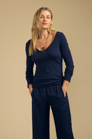 A woman with long blond hair wears the NOURA TEE by Velvet by Graham & Spencer—a navy cotton jersey top with a scoop V-neckline and long sleeves—paired with matching navy pants and a pendant necklace, standing with her hands in her pockets against a beige background.