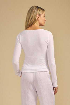 A woman is standing with her back to the camera, wearing the NOURA TEE by Velvet by Graham & Spencer—a long sleeve, scoop V-neckline white cotton jersey top—paired with loose white pants, set against a plain beige background.