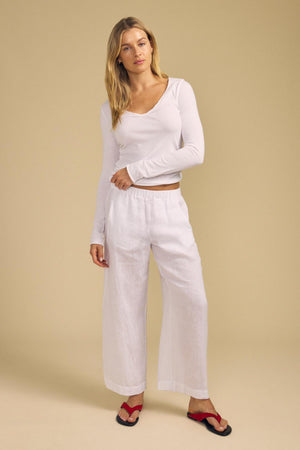 A woman stands against a beige background wearing the NOURA TEE by Velvet by Graham & Spencer—a long-sleeve white soft cotton jersey top with a scoop V-neckline—paired with loose white linen pants and red sandals.