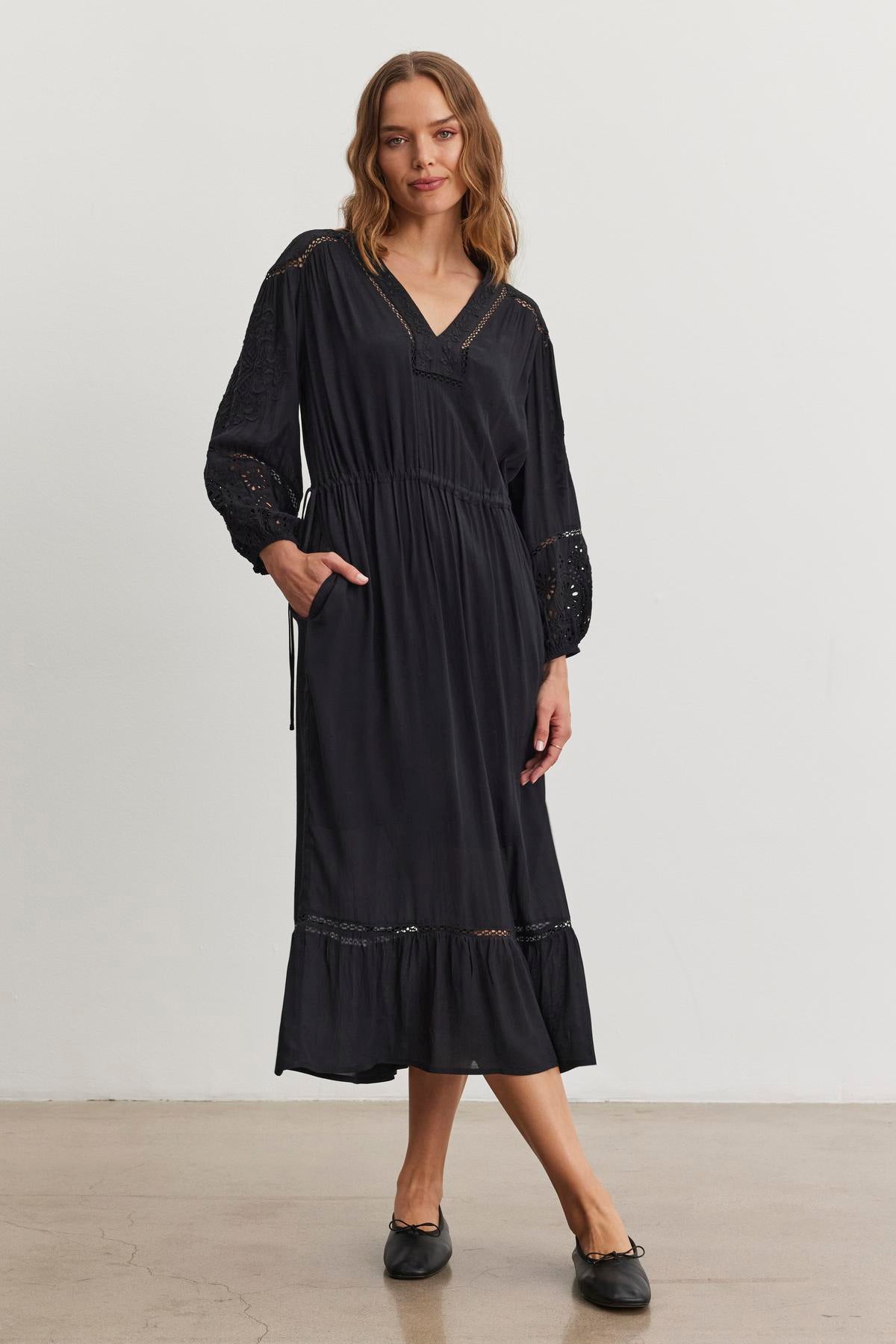   A person poses indoors against a plain background wearing the ARINA DRESS, an ankle-length black long-sleeve dress with a drawstring waist by Velvet by Graham & Spencer. 