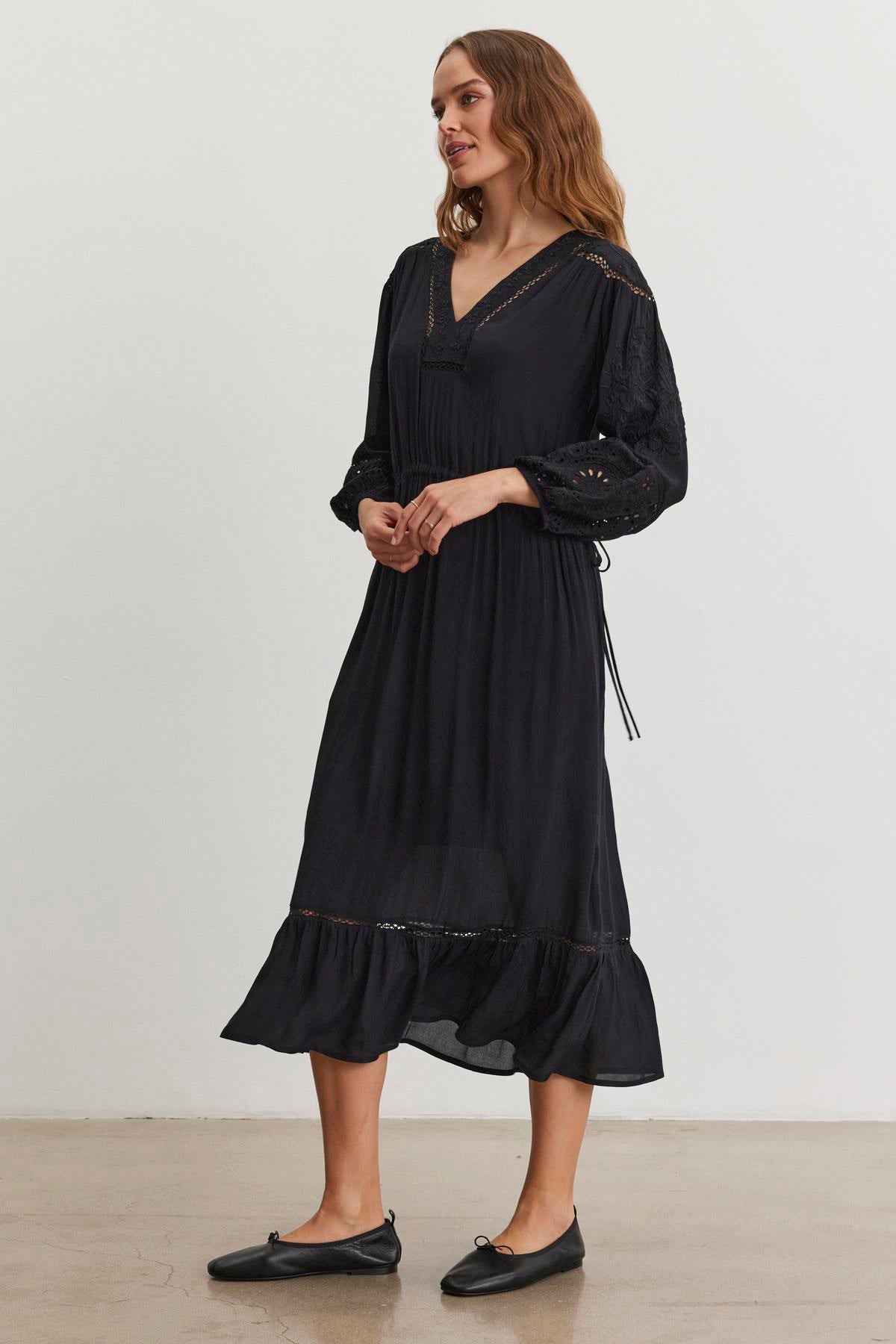   A person wearing the ARINA DRESS by Velvet by Graham & Spencer, a long-sleeved black dress with lace and embroidery, stands against a light background. With a V-neckline, they gaze to the side with their hands clasped. 