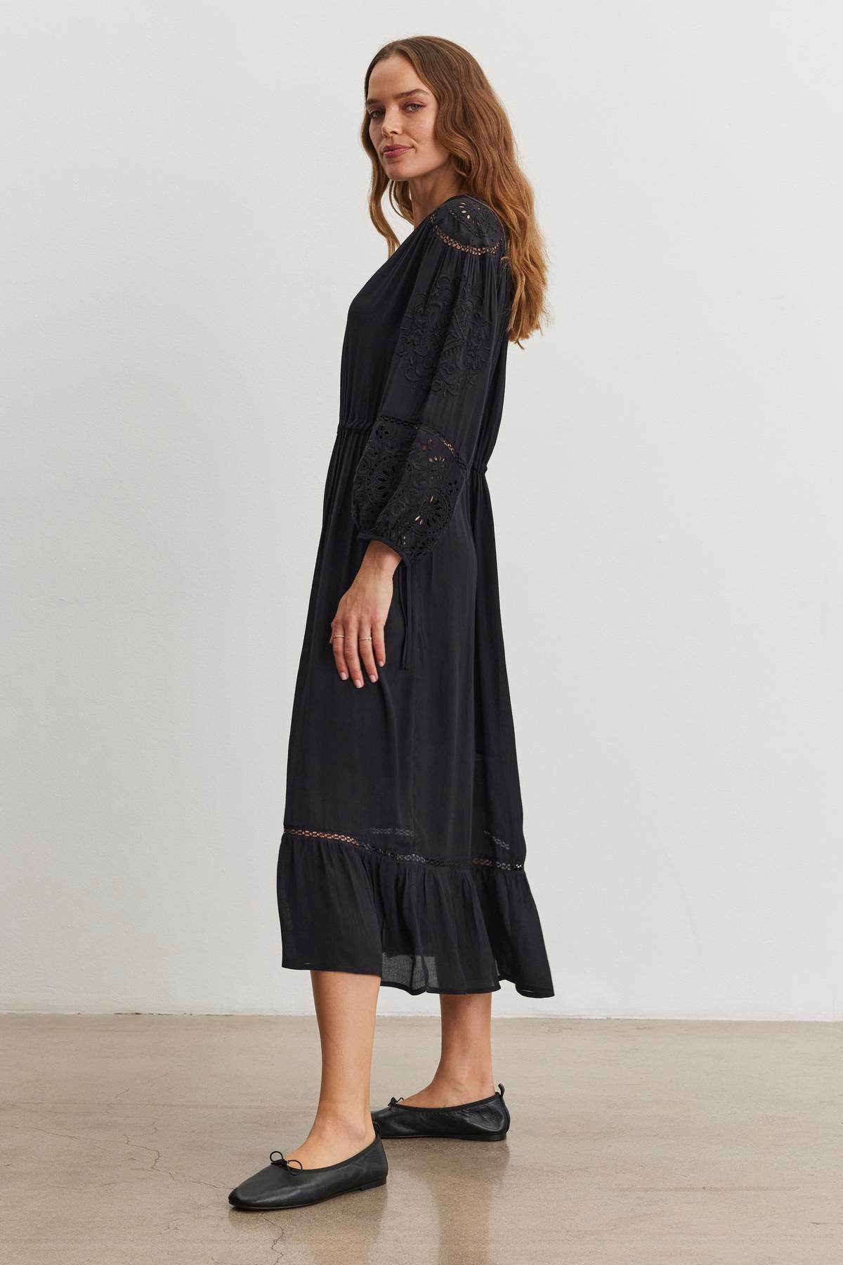   A woman with long hair stands sideways in an ARINA DRESS by Velvet by Graham & Spencer, showcasing lace details and elegant embroidery, paired with black flats on a plain background. 