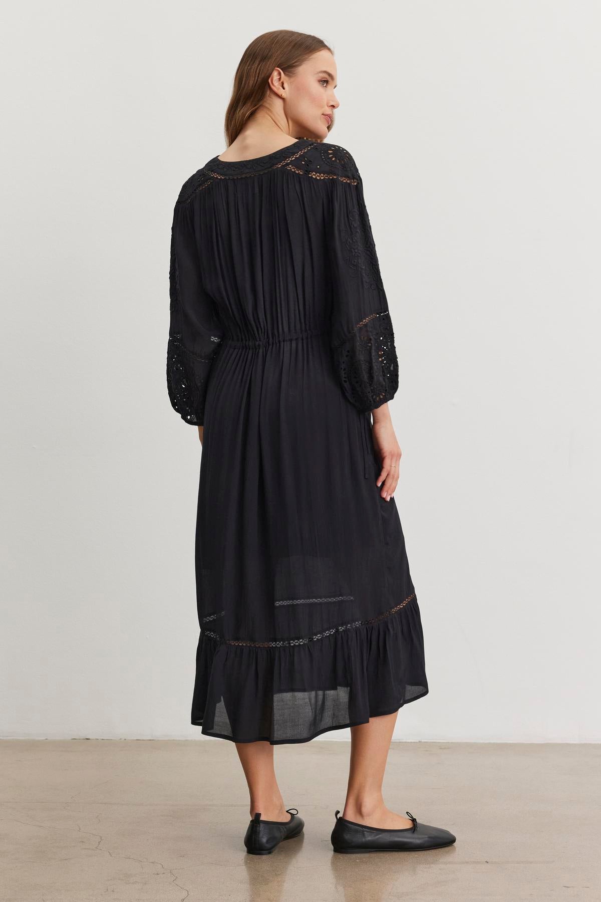   Wearing the ARINA DRESS by Velvet by Graham & Spencer, a person stands facing away. The long-sleeved black dress, featuring lace and delicate embroidery with a gathered waist, elegantly falls below the knees. Paired with black shoes, this ensemble exudes timeless grace. 