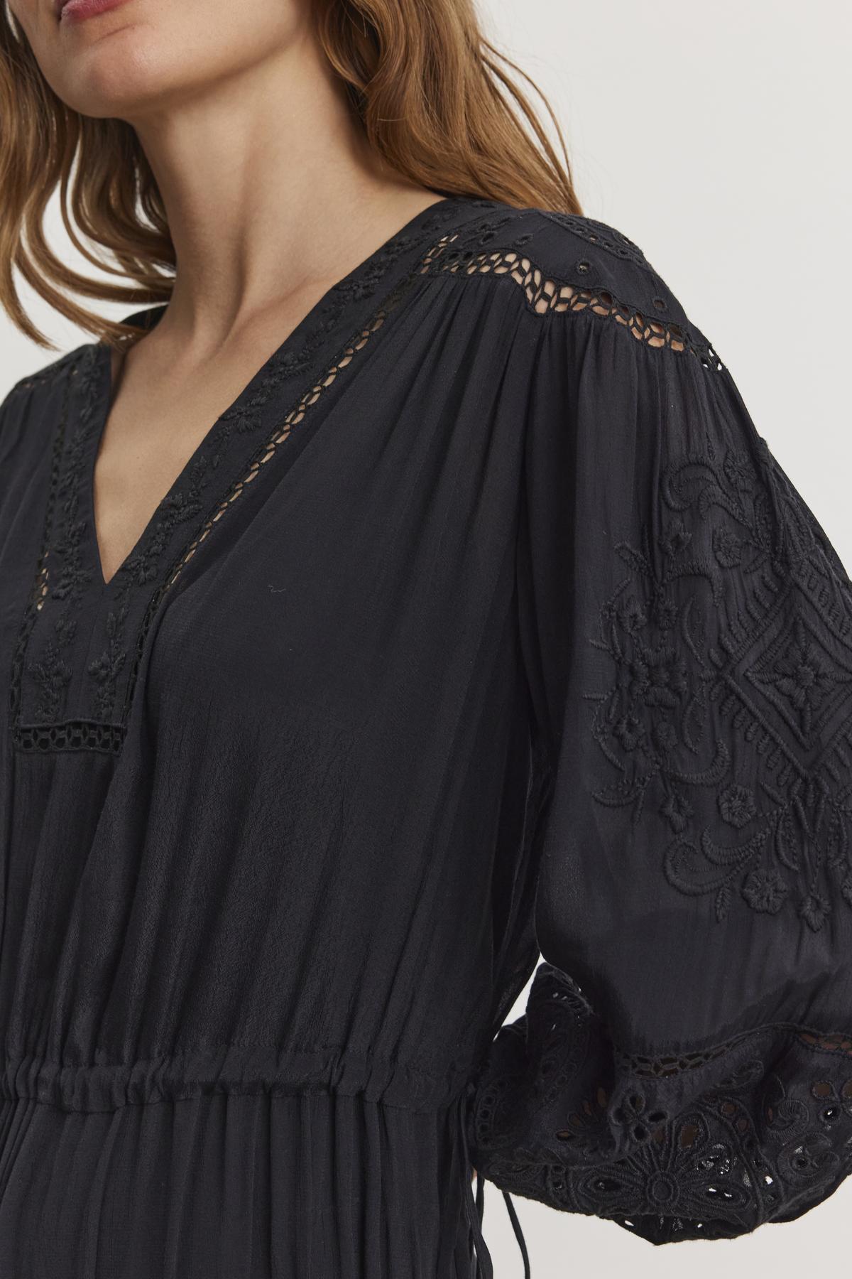   Close-up of a person wearing the ARINA DRESS by Velvet by Graham & Spencer, a black dress with intricate embroidery on the sleeves and neckline, featuring a stylish V-neckline. 