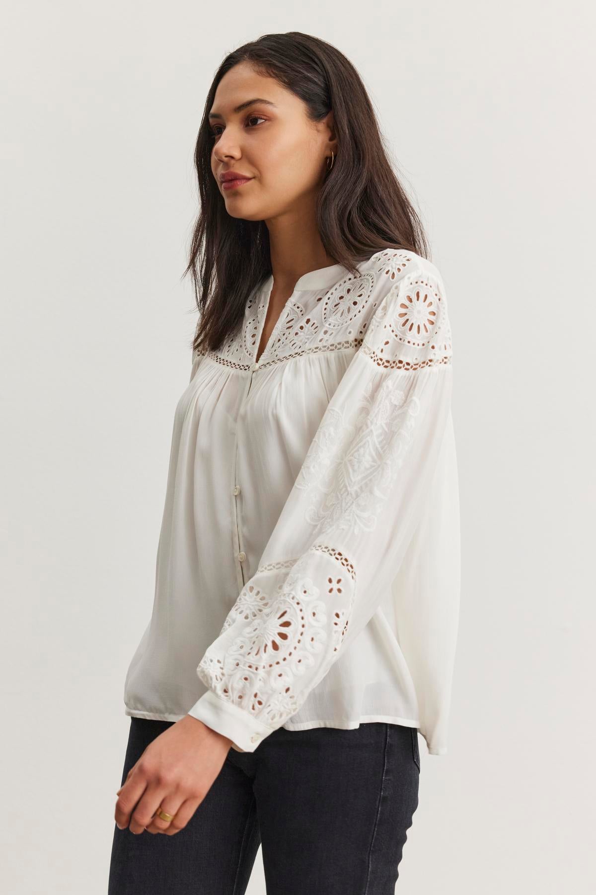   A woman wearing the LIBBY TOP from Velvet by Graham & Spencer, featuring white with eyelet embroidery, stands against a plain background. Her dark hair is loose and she's paired it with dark pants. 