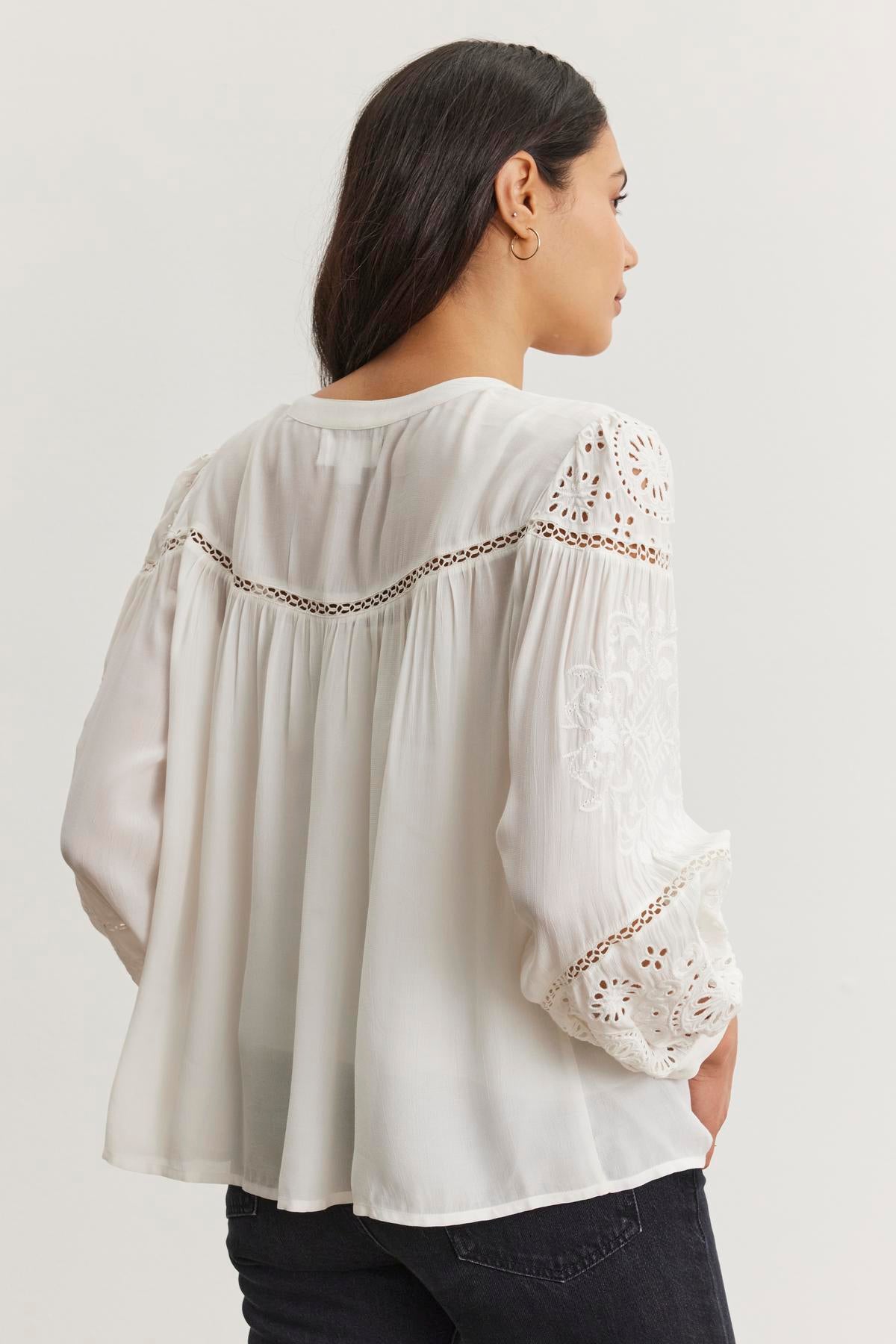   A woman models the LIBBY TOP by Velvet by Graham & Spencer, a white flowing blouse featuring eyelet and embroidery details on the button front and sleeves, against a plain background while facing away. 