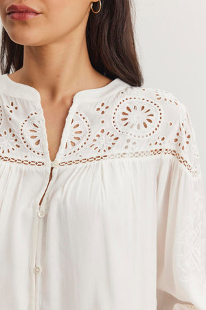 Someone is wearing the LIBBY TOP from Velvet by Graham & Spencer, a white blouse with intricate eyelet embroidery and round patterns near the neckline and shoulders.