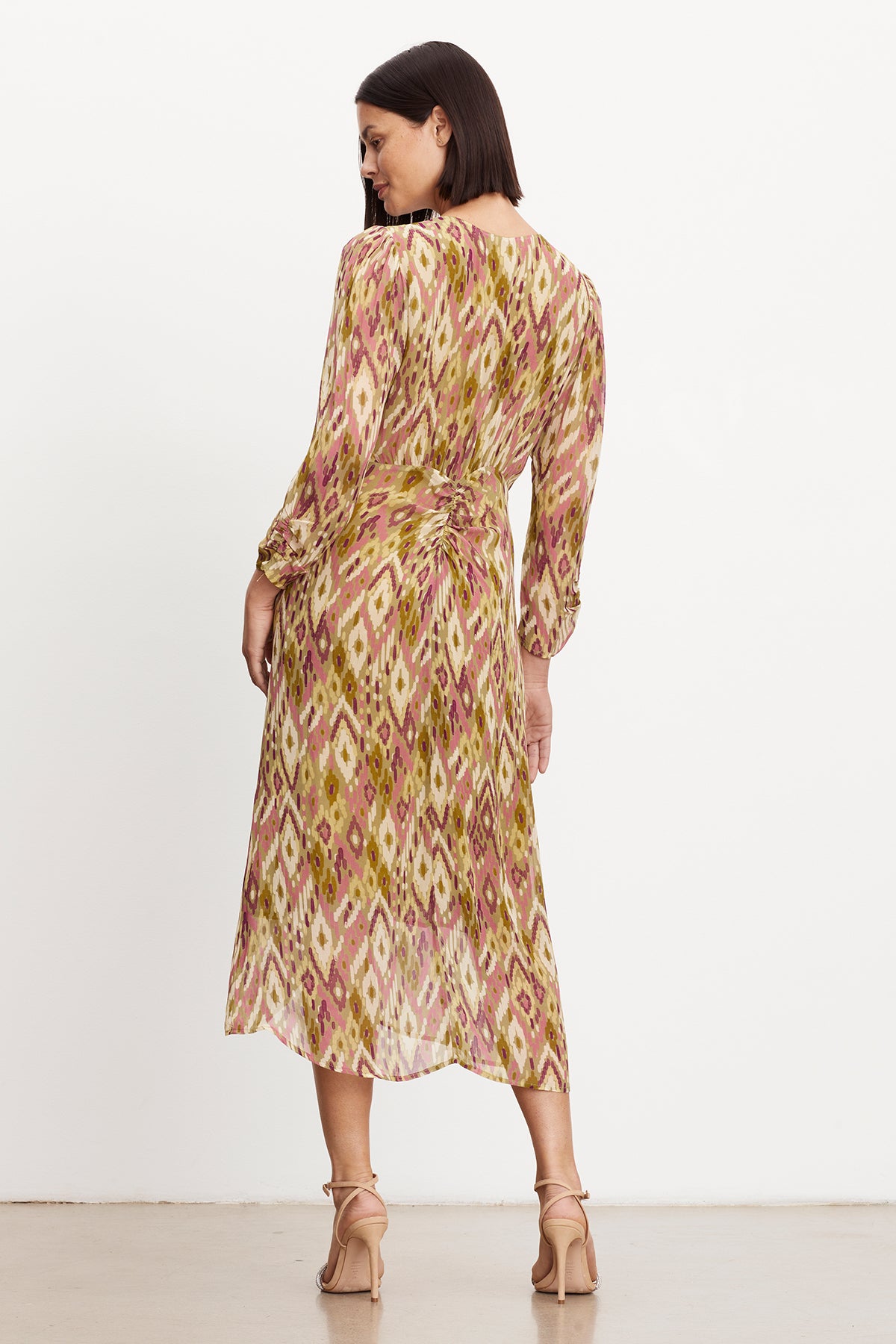 Woman standing facing away, wearing a long sleeve, mid-length CAILEY PRINTED RUCHED DRESS in viscose georgette with a pink and green bohemian print, teamed with light pink heels by Velvet by Graham & Spencer.-35577738363073