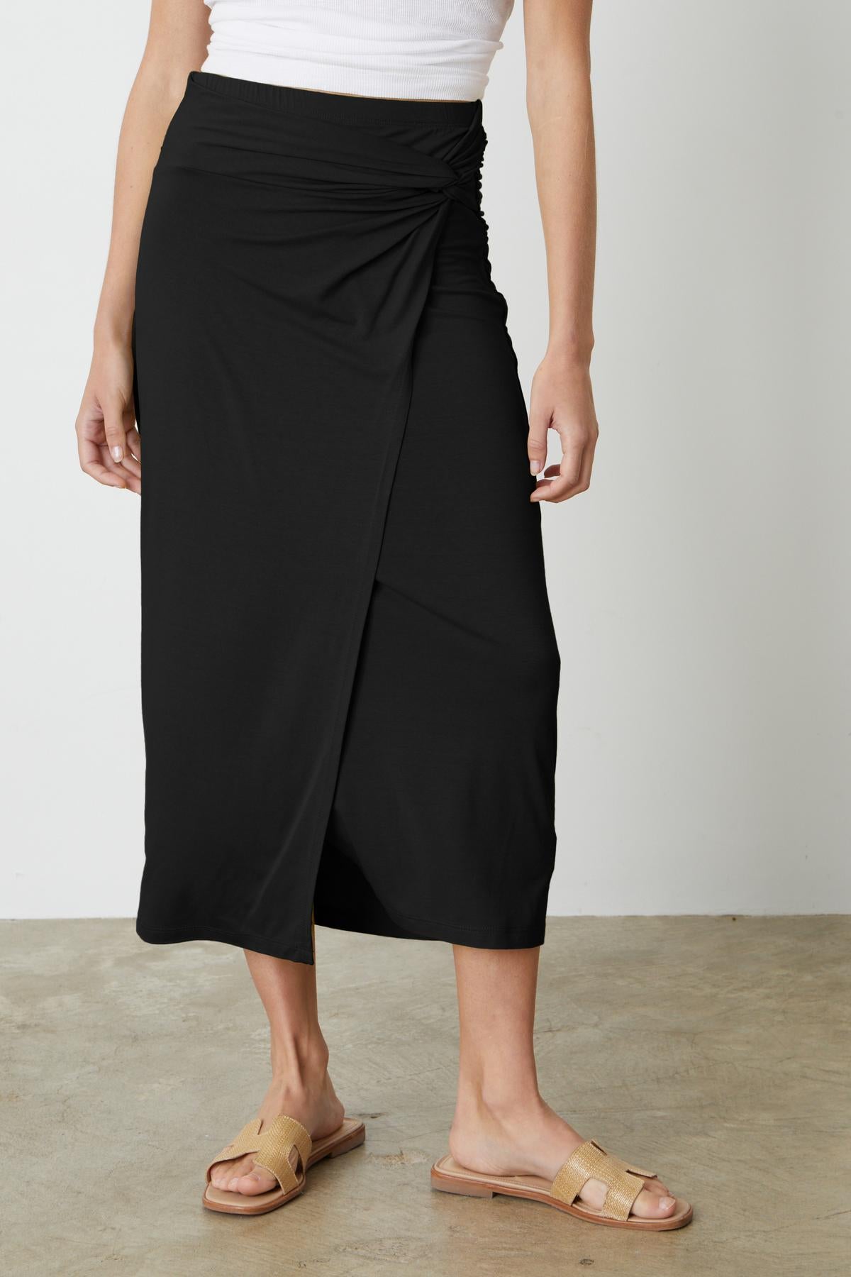 A woman wearing the Velvet by Graham & Spencer SHAE TWIST FRONT SKIRT and white sandals.-35211099799745