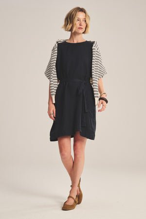 A woman wears the VIVIAN LINEN DRESS by Velvet by Graham & Spencer, a loose-fitting black mini dress with dolman sleeves and a detachable belt. She has short blond hair and brown flats.