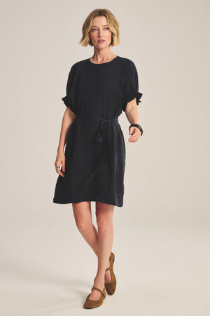 A woman stands against a plain light background wearing the VIVIAN LINEN DRESS by Velvet by Graham & Spencer—a woven linen mini dress with dolman sleeves, a waist tie, and brown flat shoes.