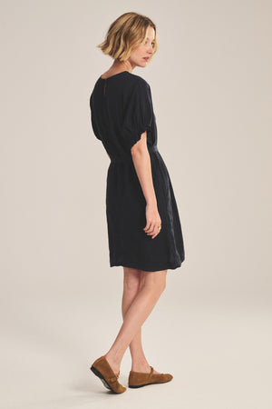 A woman poses sideways against a light background, wearing the VIVIAN LINEN DRESS by Velvet by Graham & Spencer—a short-sleeve linen mini dress with a detachable belt—paired with brown flat shoes.