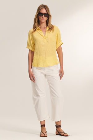 A person wearing the VIVICA LINEN TOP by Velvet by Graham & Spencer, paired with white wide-leg pants, brown sandals, sunglasses, and a pearl necklace stands against a plain background.