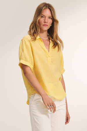 A woman stands against a plain light background, wearing the Velvet by Graham & Spencer VIVICA LINEN TOP in yellow with short sleeves and loose fit, paired with white pants.