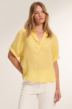 A woman wears the VIVICA LINEN TOP by Velvet by Graham & Spencer—a light yellow short-sleeve blouse with rolled cuffs—paired with white pants and a pearl necklace, standing against a plain light background.