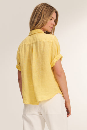 A woman faces away, wearing the VIVICA LINEN TOP by Velvet by Graham & Spencer—a short-sleeved yellow button-up shirt—paired with white pants against a plain background.