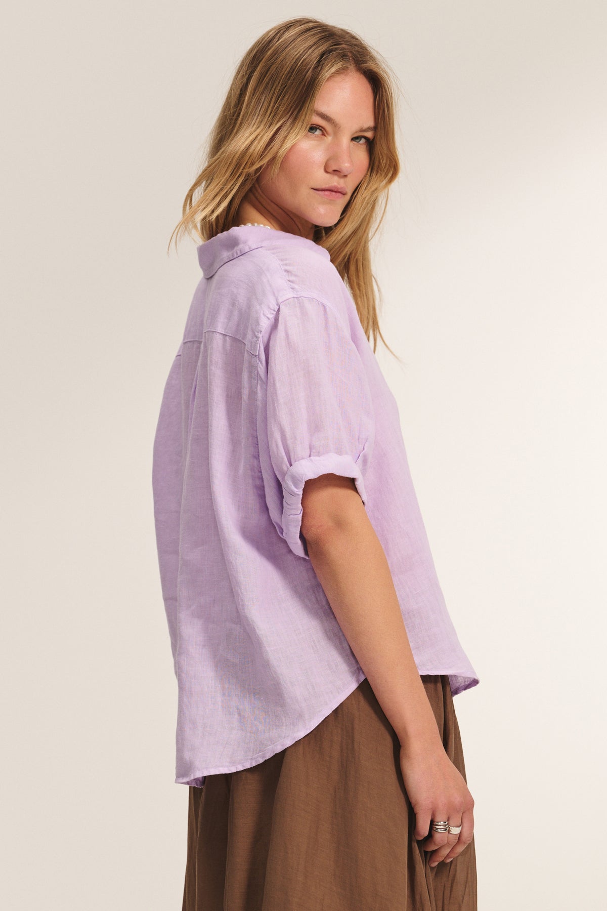A woman stands sideways against a plain background, wearing the VIVICA LINEN TOP by Velvet by Graham & Spencer—a light purple, short-sleeve button-up shirt—paired with a brown skirt.-39173165154497