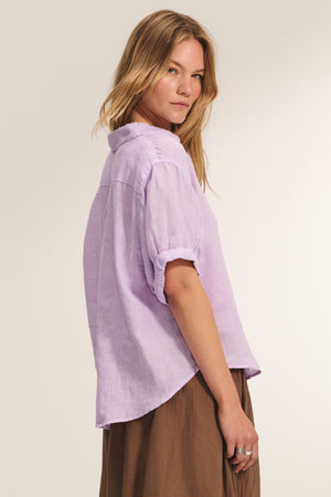 A woman stands sideways against a plain background, wearing the VIVICA LINEN TOP by Velvet by Graham & Spencer—a light purple, short-sleeve button-up shirt—paired with a brown skirt.