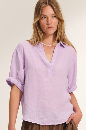 A woman wearing the VIVICA LINEN TOP by Velvet by Graham & Spencer—a light purple, loose-fitting linen shirt with rolled sleeves—paired with a pearl necklace, stands against a plain background.