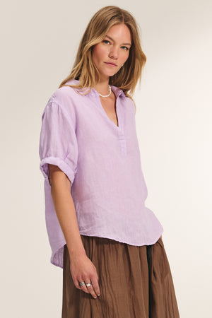 A woman wearing the VIVICA LINEN TOP by Velvet by Graham & Spencer, a light purple long-sleeve blouse with rolled sleeves, paired with a brown skirt, stands against a plain light background.