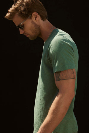 A man in a stylish green AMARO TEE by Velvet by Graham & Spencer and sunglasses is standing against a dark background, looking down with a neutral expression. He has short hair and a tattoo on his upper arm.