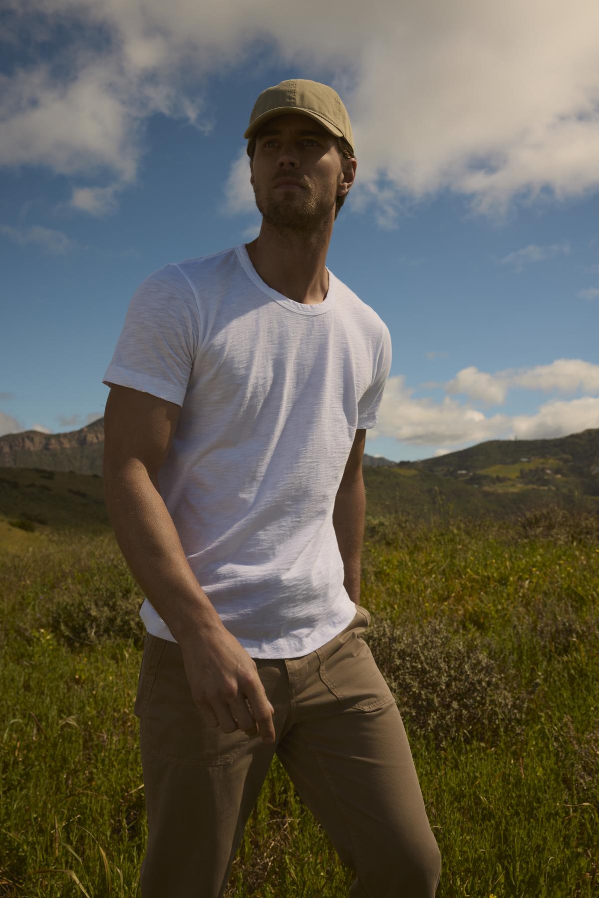   A man in a white Velvet by Graham & Spencer t-shirt and khaki pants stands in a field with a picturesque, cloudy sky above and mountains in the distance. He is wearing a tailored jacket and a cap, looking away. 
