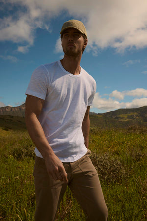 A man in a white Velvet by Graham & Spencer t-shirt and khaki pants stands in a field with a picturesque, cloudy sky above and mountains in the distance. He is wearing a tailored jacket and a cap, looking away.