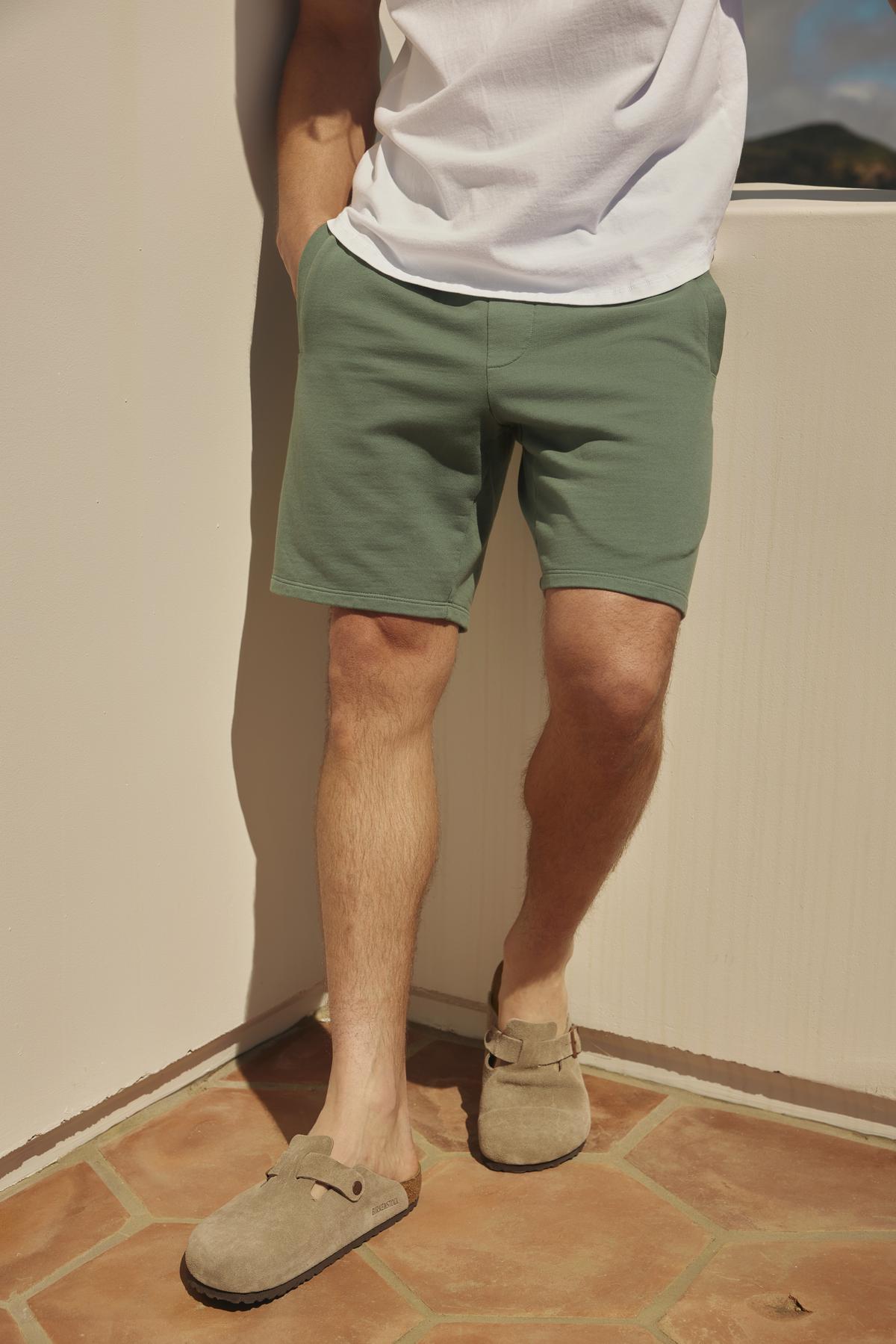 Man standing on a tiled patio wearing Velvet by Graham & Spencer's BECKETT SHORTs, a white t-shirt, and gray loafers, facing a sunlit wall.-36753585766593