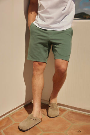 Man standing on a tiled patio wearing Velvet by Graham & Spencer's BECKETT SHORTs, a white t-shirt, and gray loafers, facing a sunlit wall.
