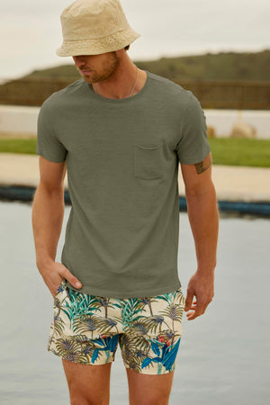 A man wearing a beige bucket hat, a gray CHAD TEE by Velvet by Graham & Spencer with raw edge details, and floral-patterned shorts stands near a body of water with greenery in the background.