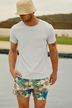 A man wearing a beige bucket hat, a white CHAD TEE by Velvet by Graham & Spencer with a classic crew neckline, and tropical patterned shorts stands near a body of water with a blurred background of greenery.