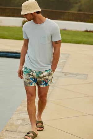 Man in a bucket hat, Velvet by Graham & Spencer CHAD TEE with raw-edge details, and tropical print shorts walking by a poolside.