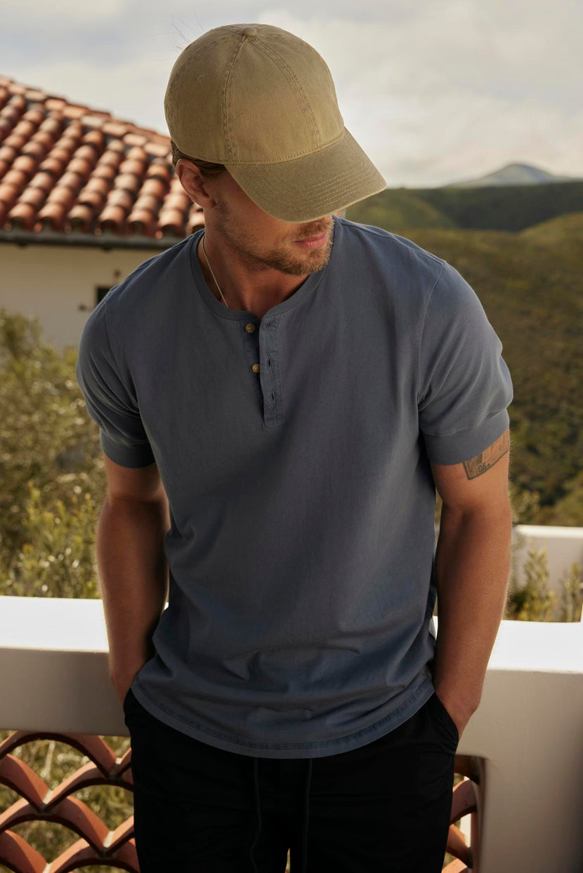 A man in a cap and DEON HENLEY t-shirt gazes downward, standing on a balcony with scenic hills and a tiled roof in the background.