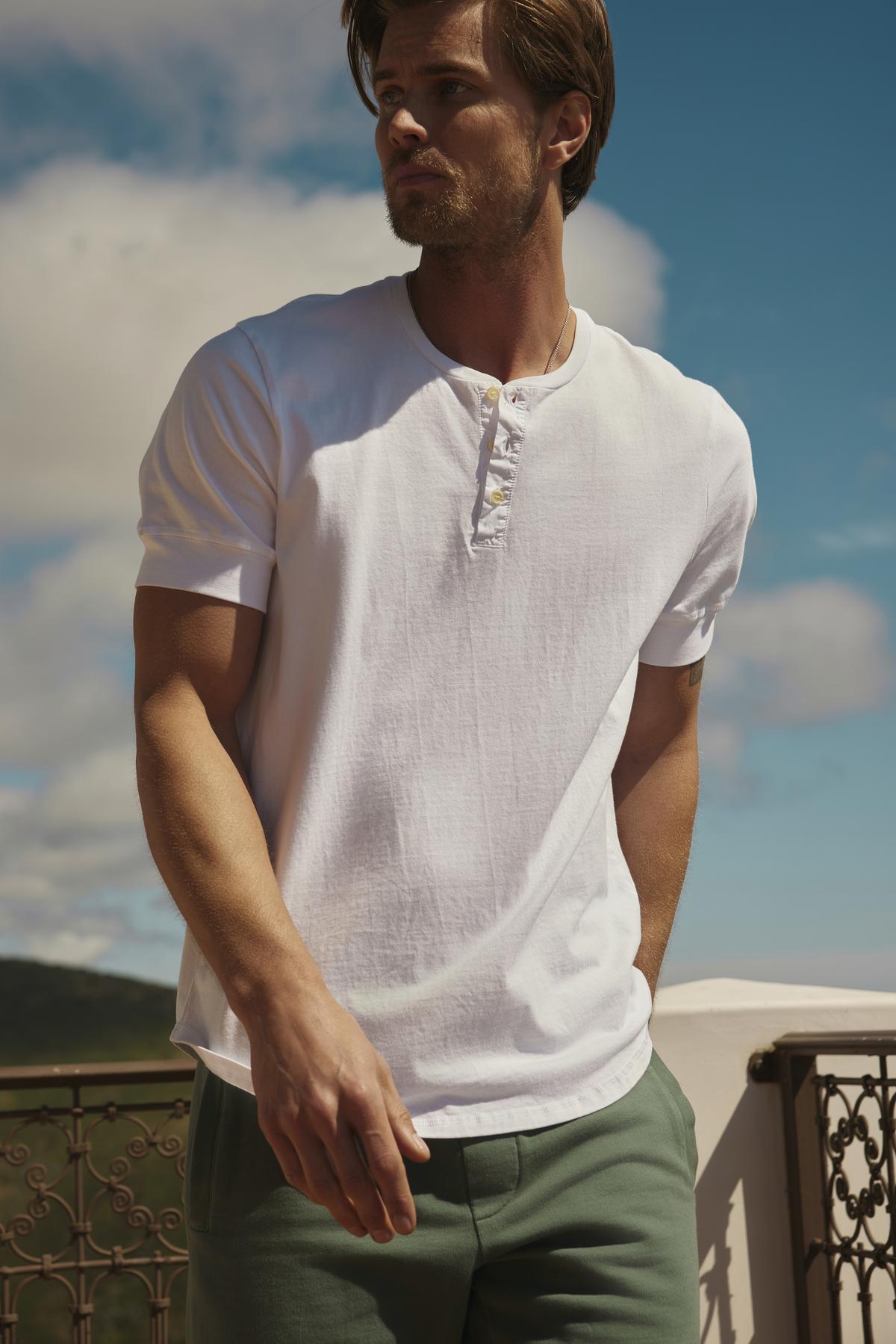   A man in a white Velvet by Graham & Spencer DEON HENLEY shirt with scooped hem and green trousers stands on a balcony, looking to the side with a backdrop of hills and sky. 